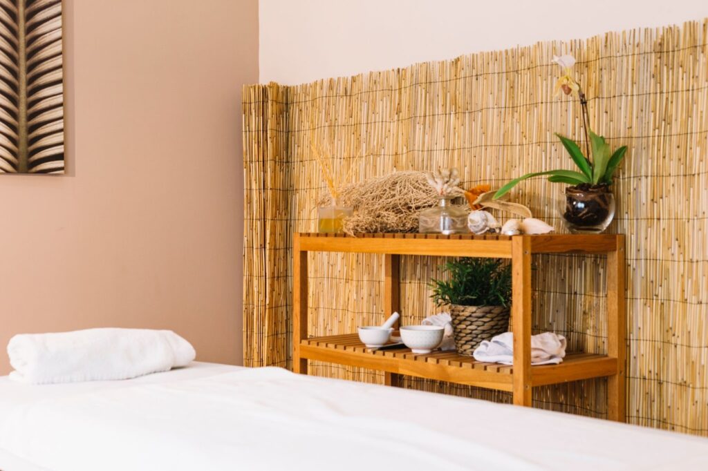 Spa treatment room with massage bed, bamboo wall decor, wooden shelves, and neatly arranged towels